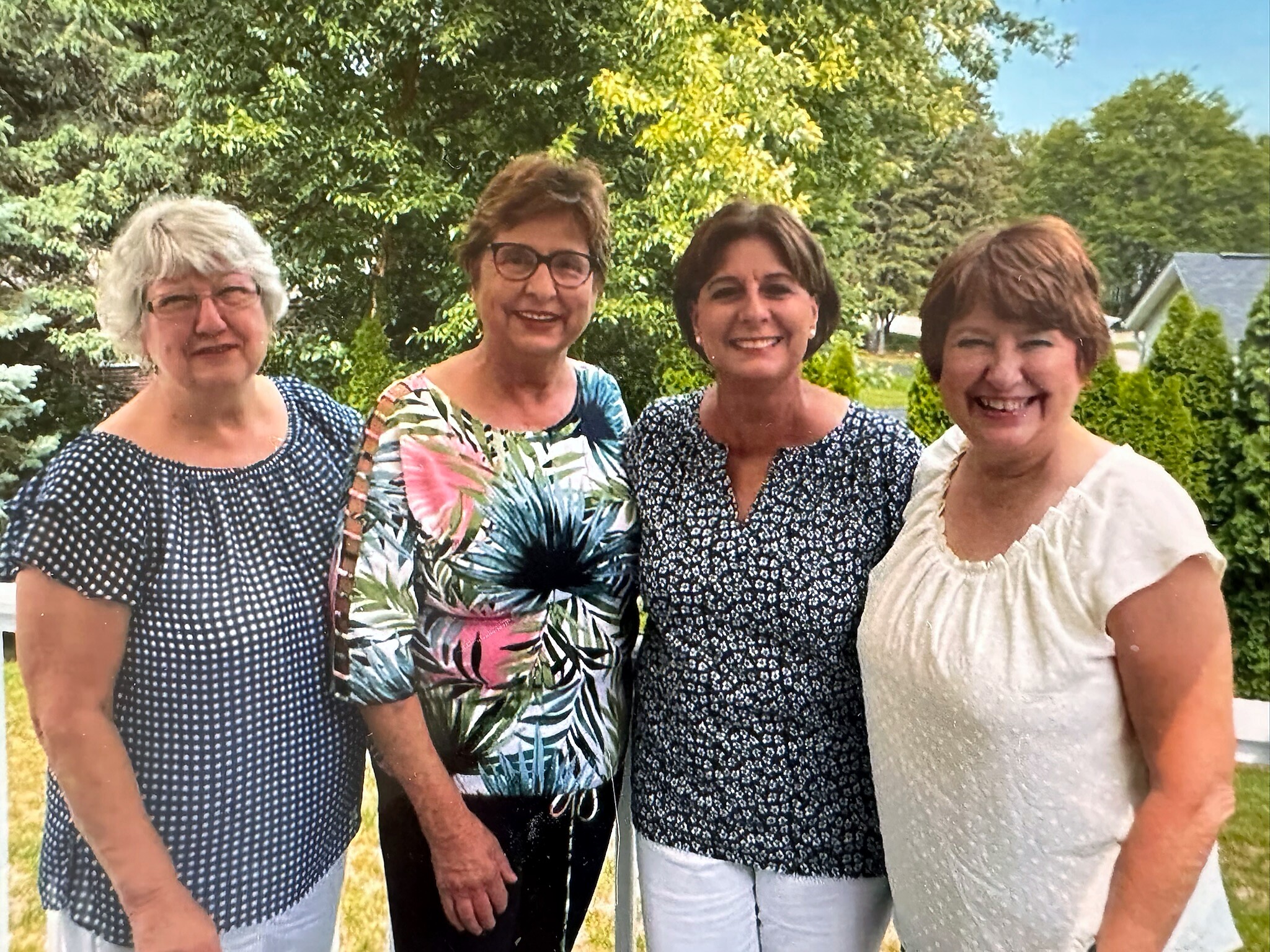 Four smiling women posing together outdoors, shown from mid‑torso up, in a leafy garden setting with a blue sky behind them.