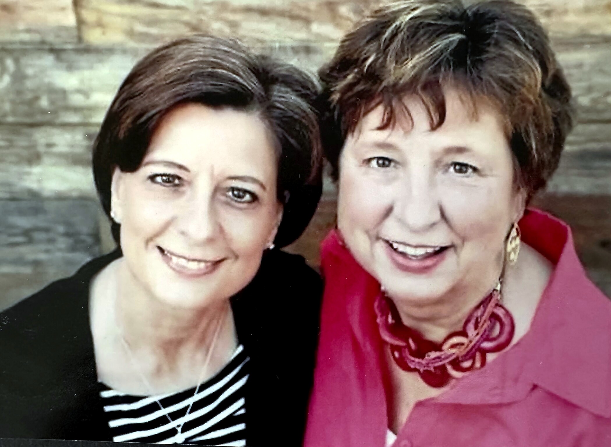 Two smiling women posing close together for a photo, one in black and white stripes and the other in a pink top with a scarf.
