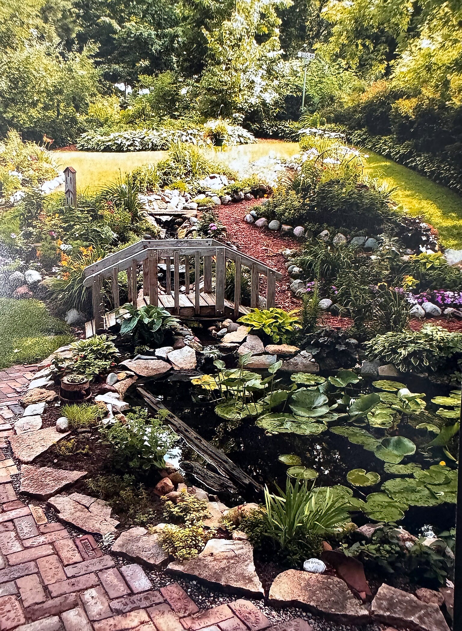 Wooden footbridge crosses a koi pond with lily pads in a lush garden setting.