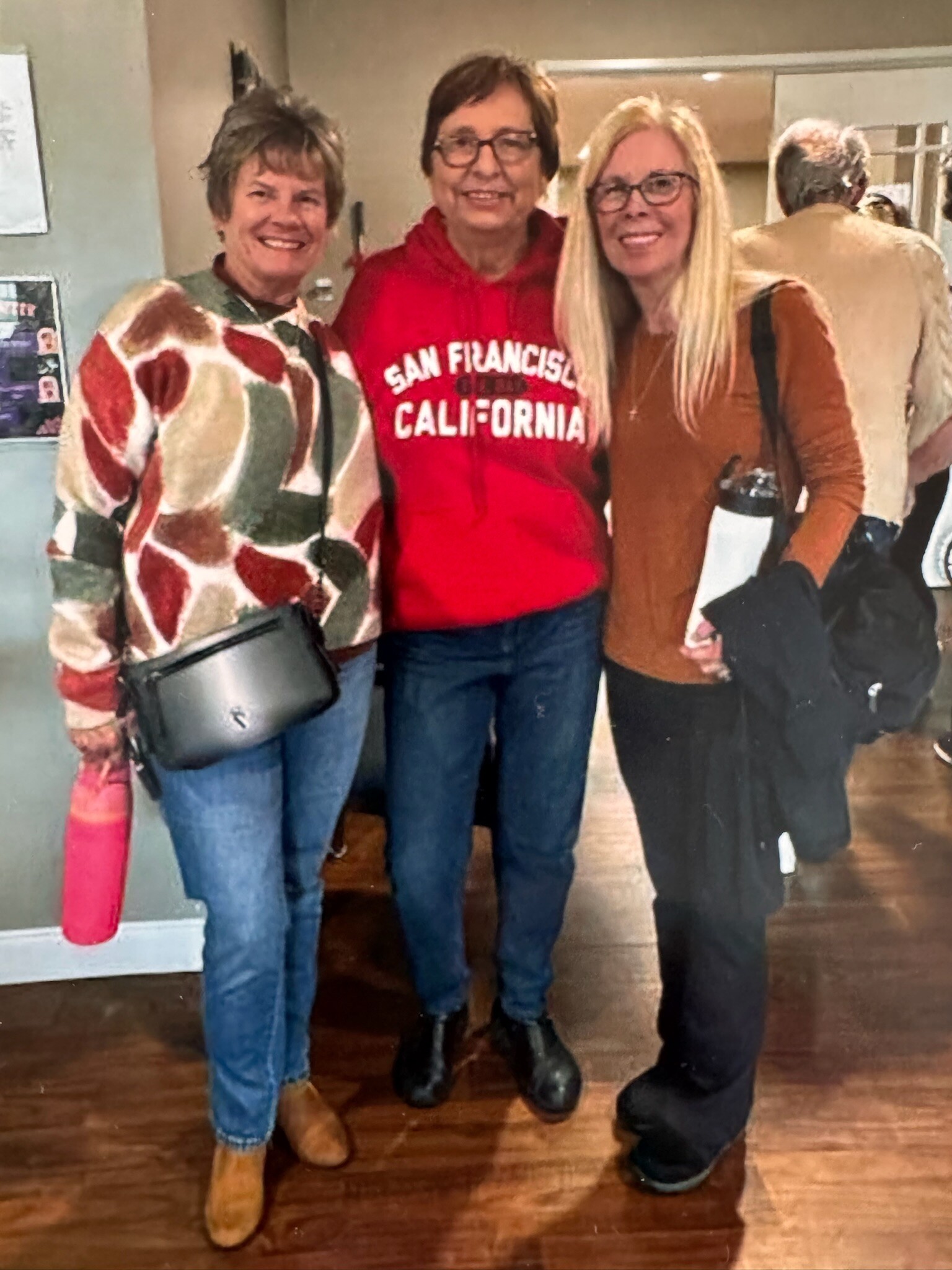 Three women stand together in a hallway, smiling at the camera: left in a colorful knit sweater with a black crossbody bag, middle in a red San Francisco hoodie, right in a brown long-sleeve top with glasses and blonde hair.