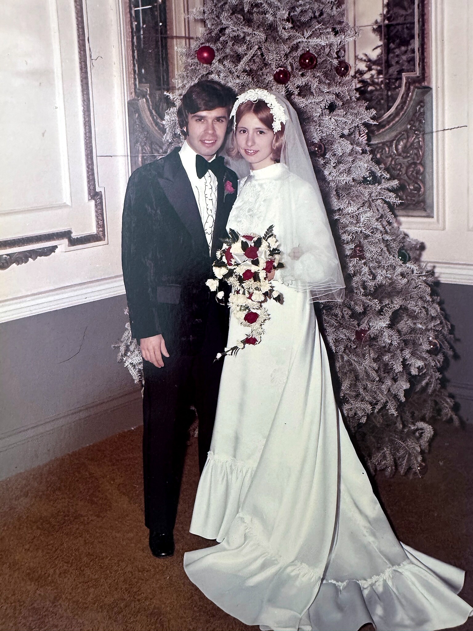 Bride and groom in formal wedding attire pose together indoors, with a frosted Christmas tree behind them and the bride holding a white-and-rose bouquet.