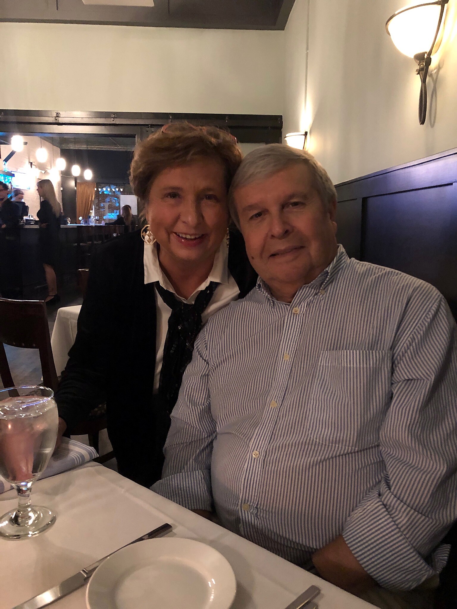 Smiling couple sits at a restaurant table; woman on left wears a black jacket and scarf, man on right wears a blue striped shirt.