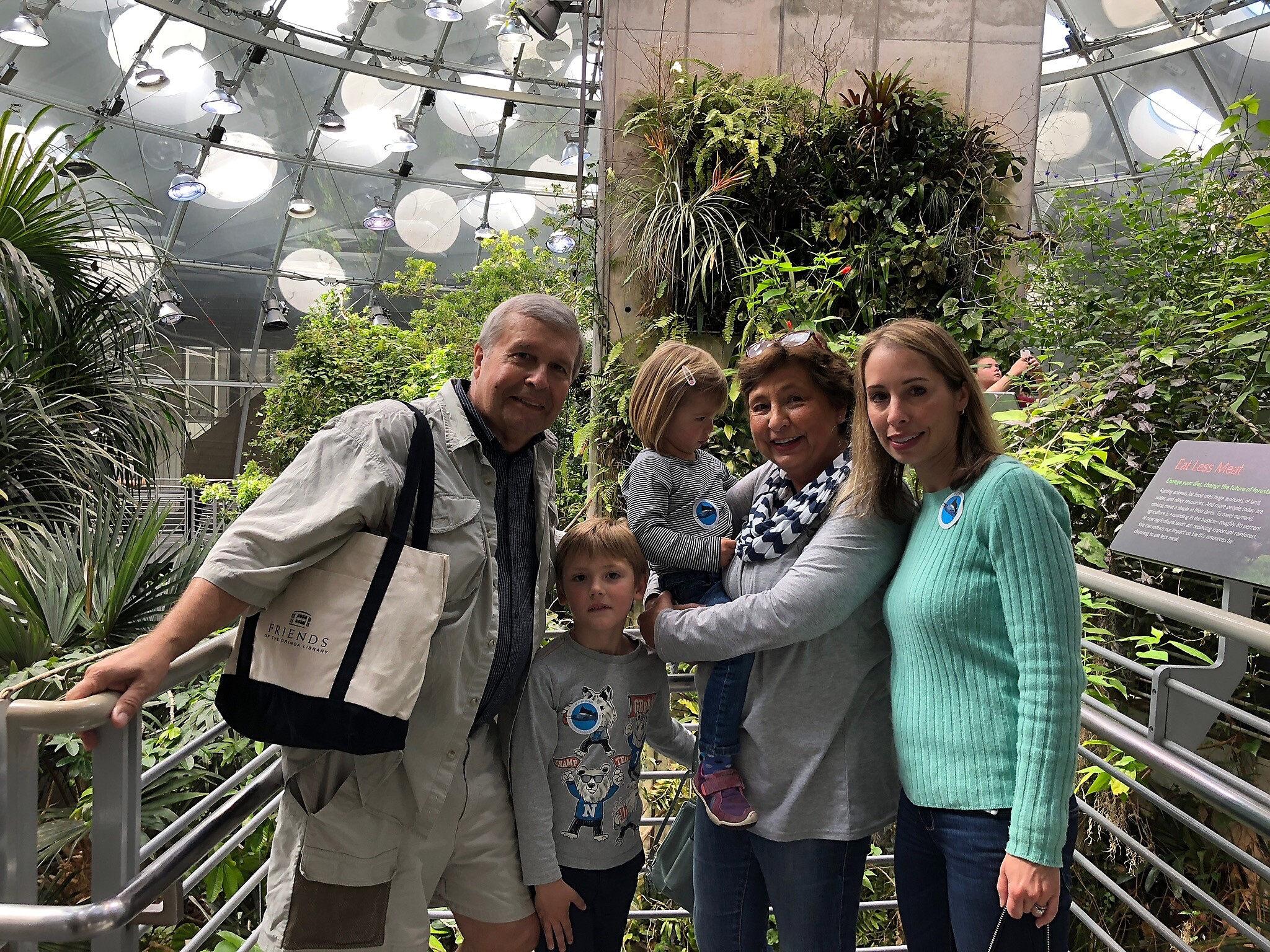 Group of five people posing on a metal railing in a lush indoor greenhouse. (two adults, two children, one younger child)