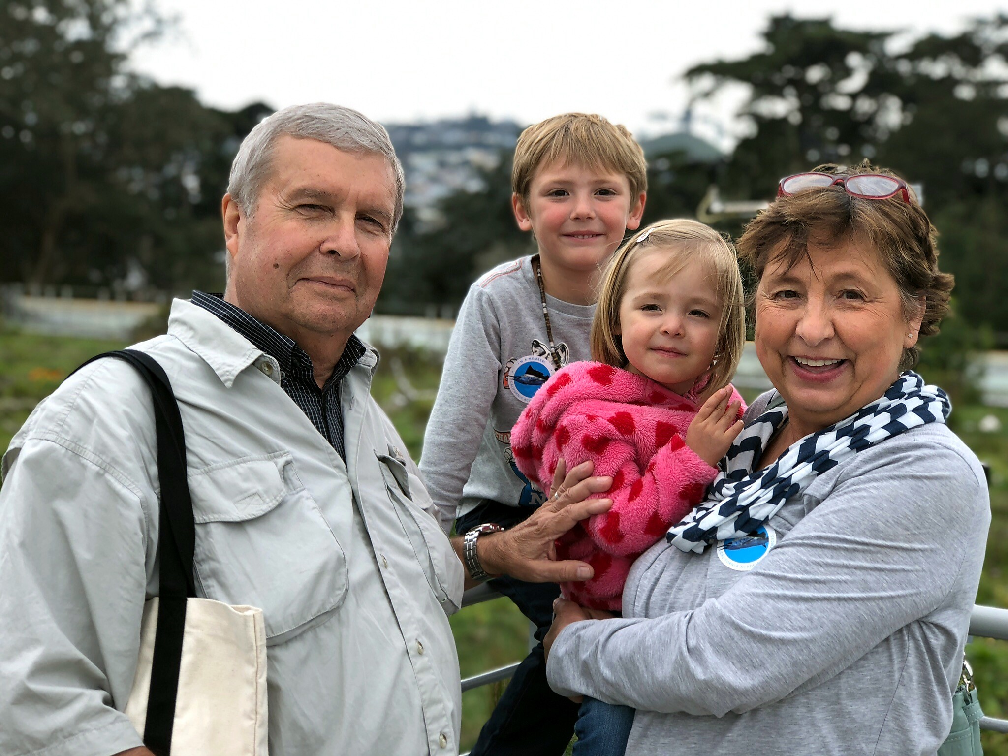 Family posing outdoors: smiling grandparents with two young children in a sunny park setting, near a railing and greenery.