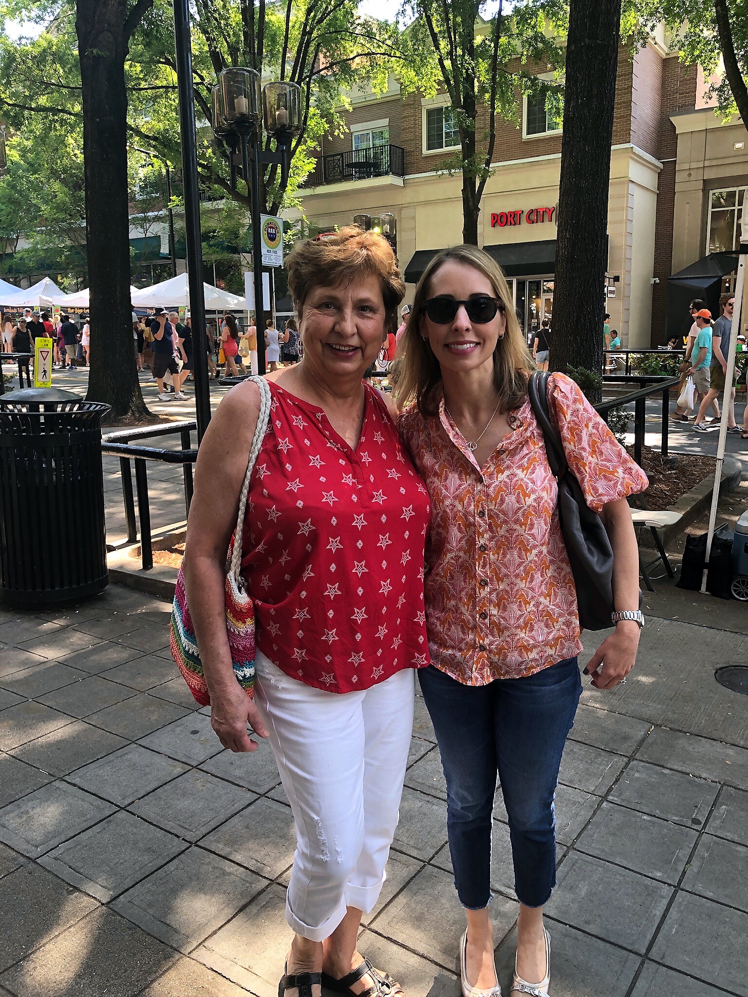 Two smiling women posing together outdoors on a sunny street market with stalls behind them and storefronts in the background.