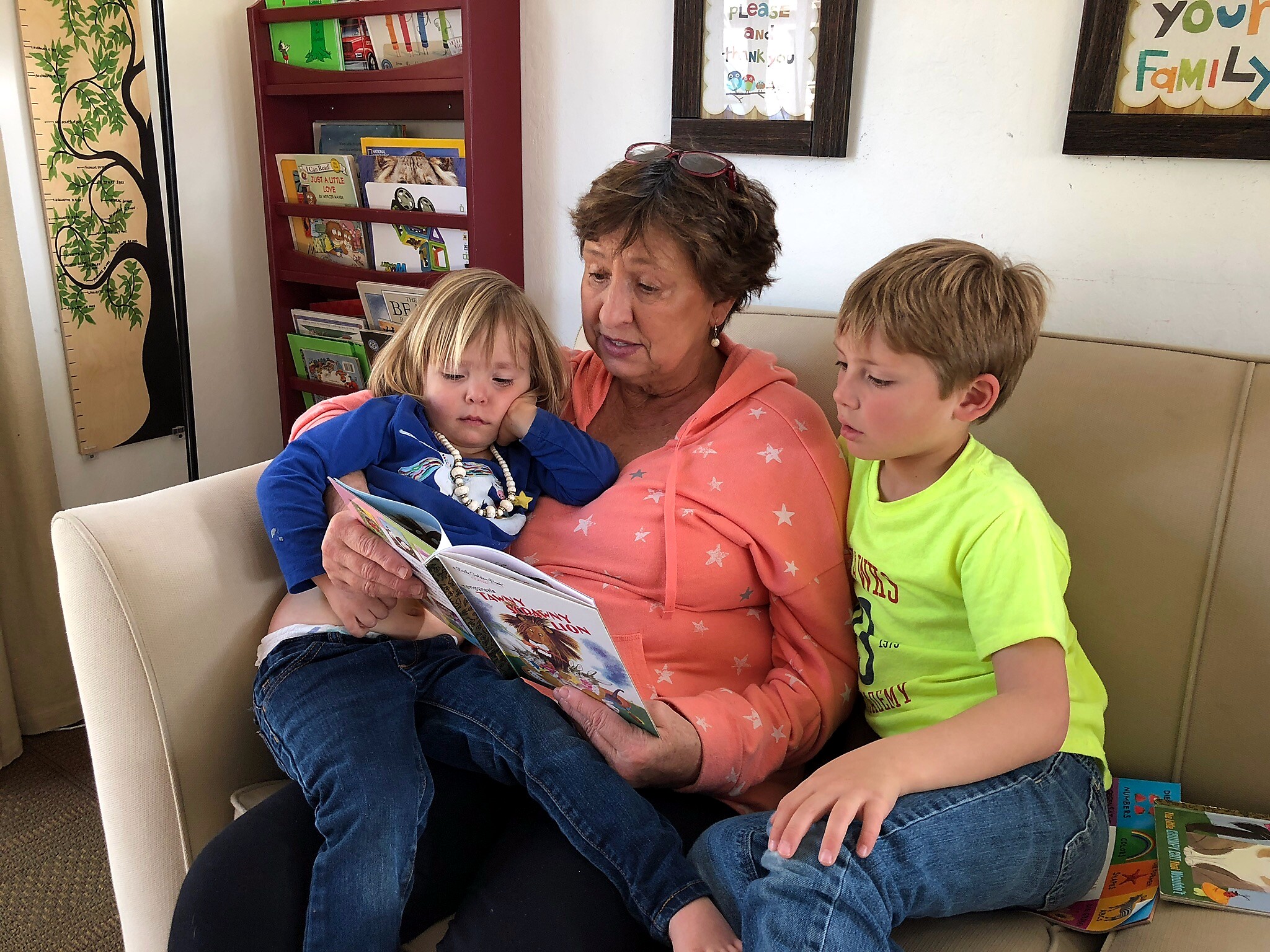 Adult woman reading a colorful children's book to two kids cuddled on a beige sofa, in a cozy reading corner.