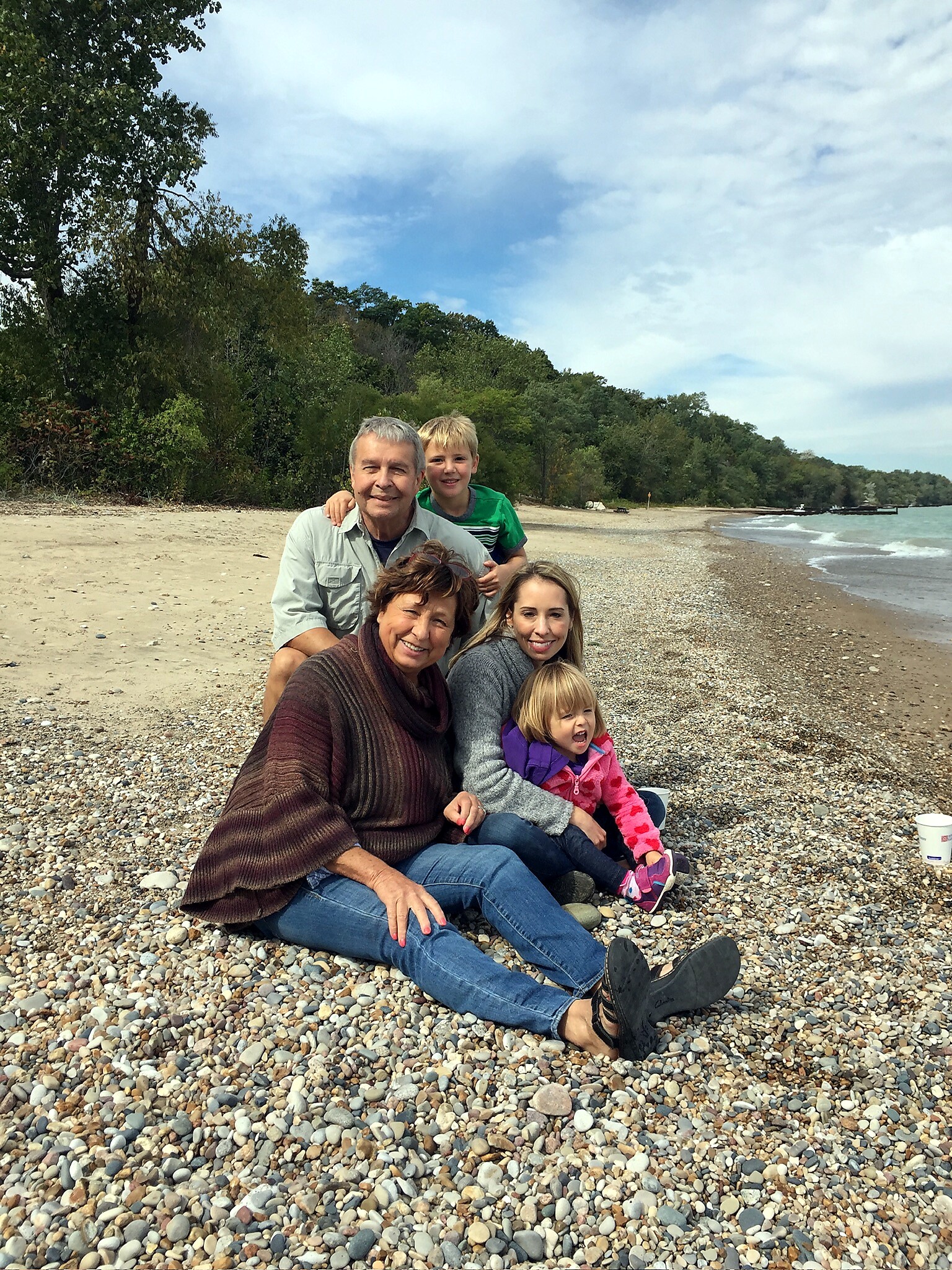 A smiling family sits together on a pebbled beach with trees behind and waves on the right side of the shore.