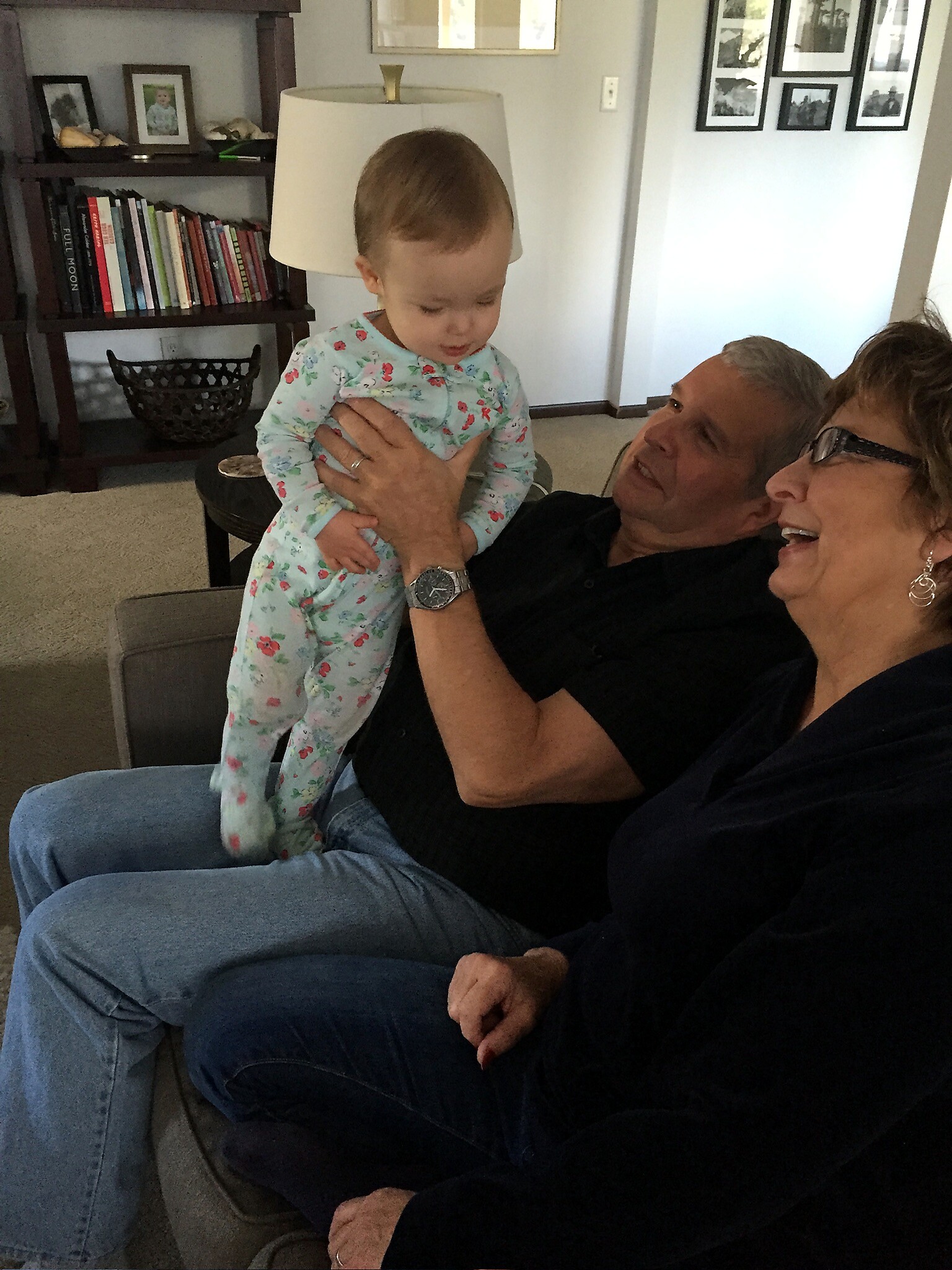 Grandfather holding a baby girl in a floral onesie while grandmother smiles on a sofa in a living room with bookshelves in the background.