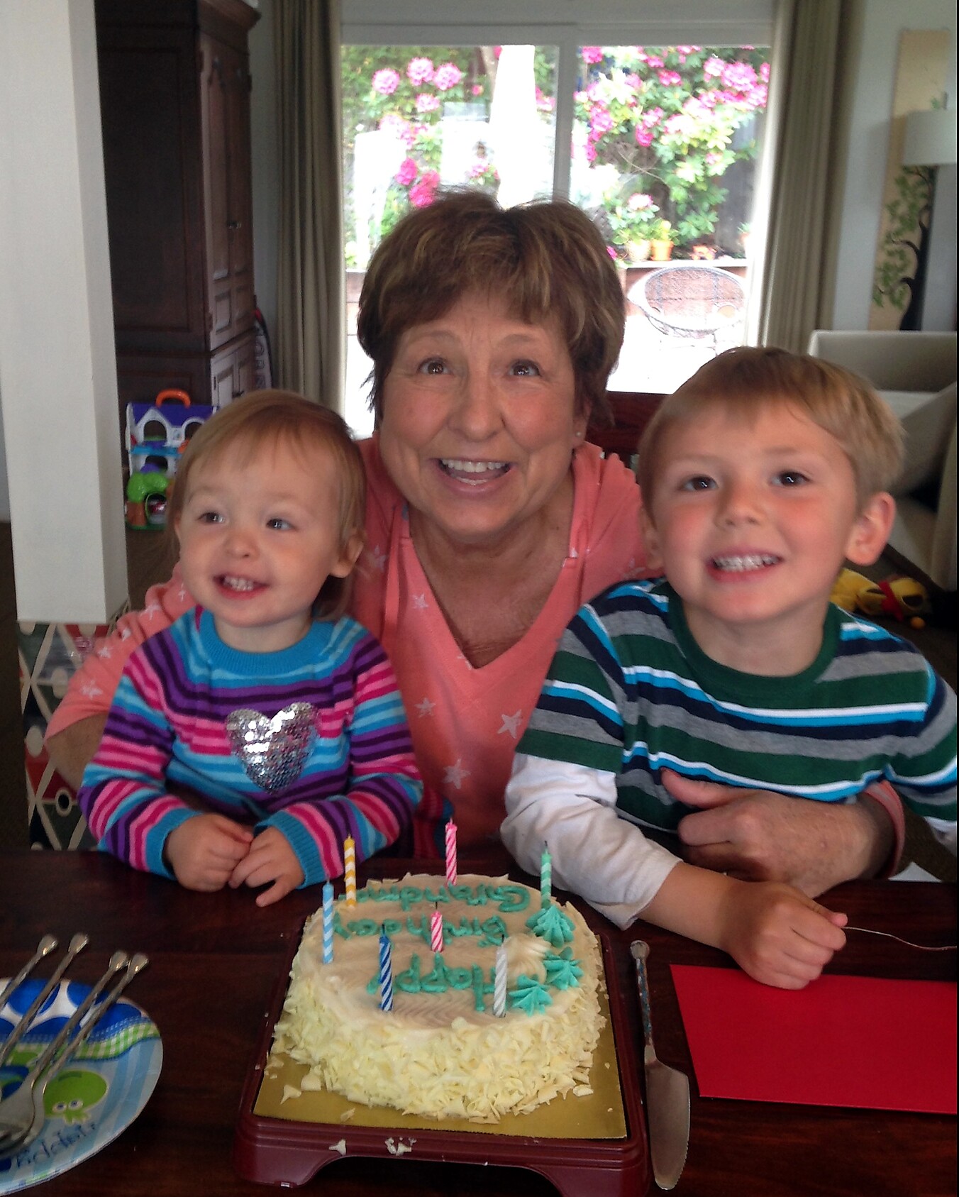 Grandmother smiles with two young children at a birthday table, a cake with lit candles in front of them.