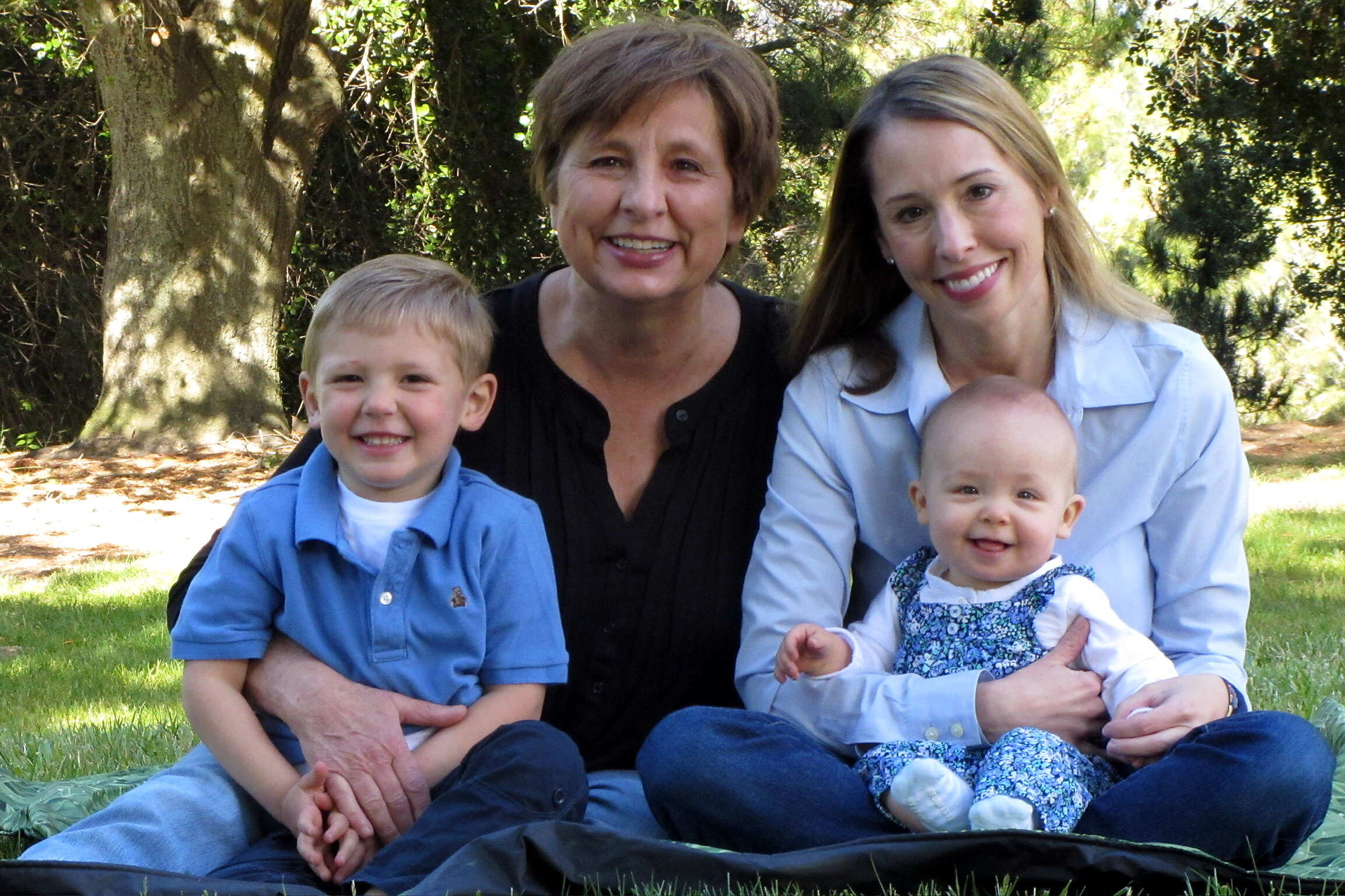 Smiling family portrait outdoors on grass with trees in the background: a young boy on the left, a middle-aged woman in the center, and a smiling woman with a baby on the right.