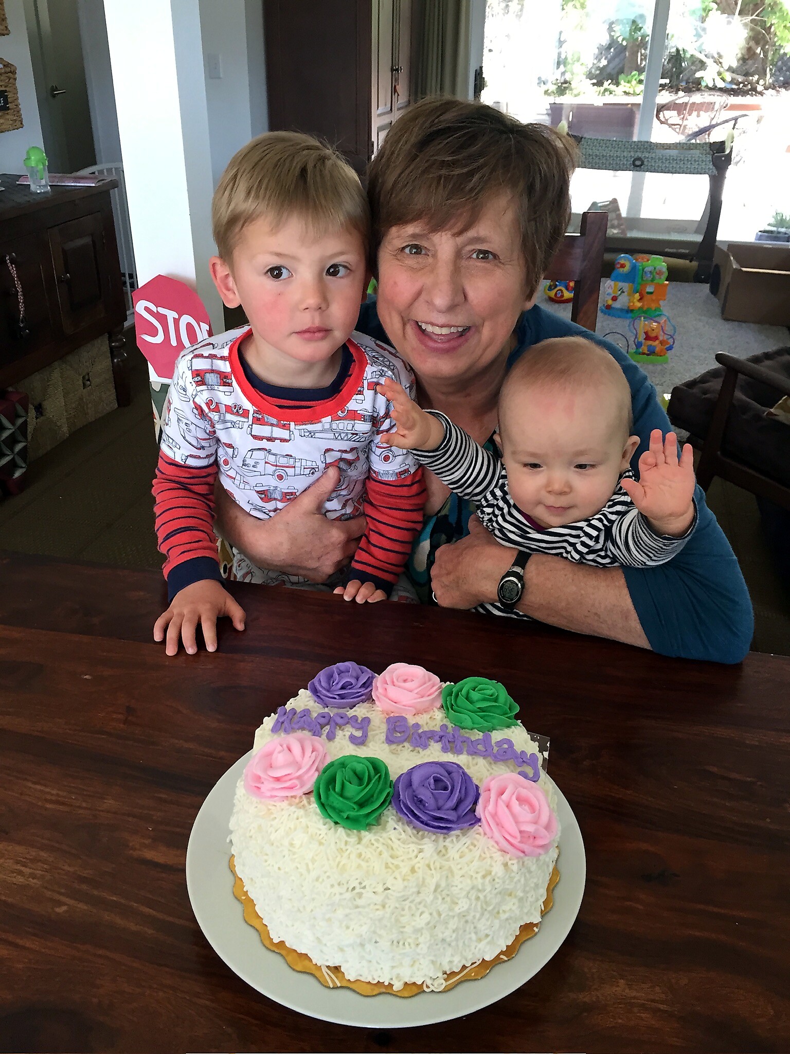Smiling grandmother holding two young children at a birthday party with a frosted cake in front of them on a table.