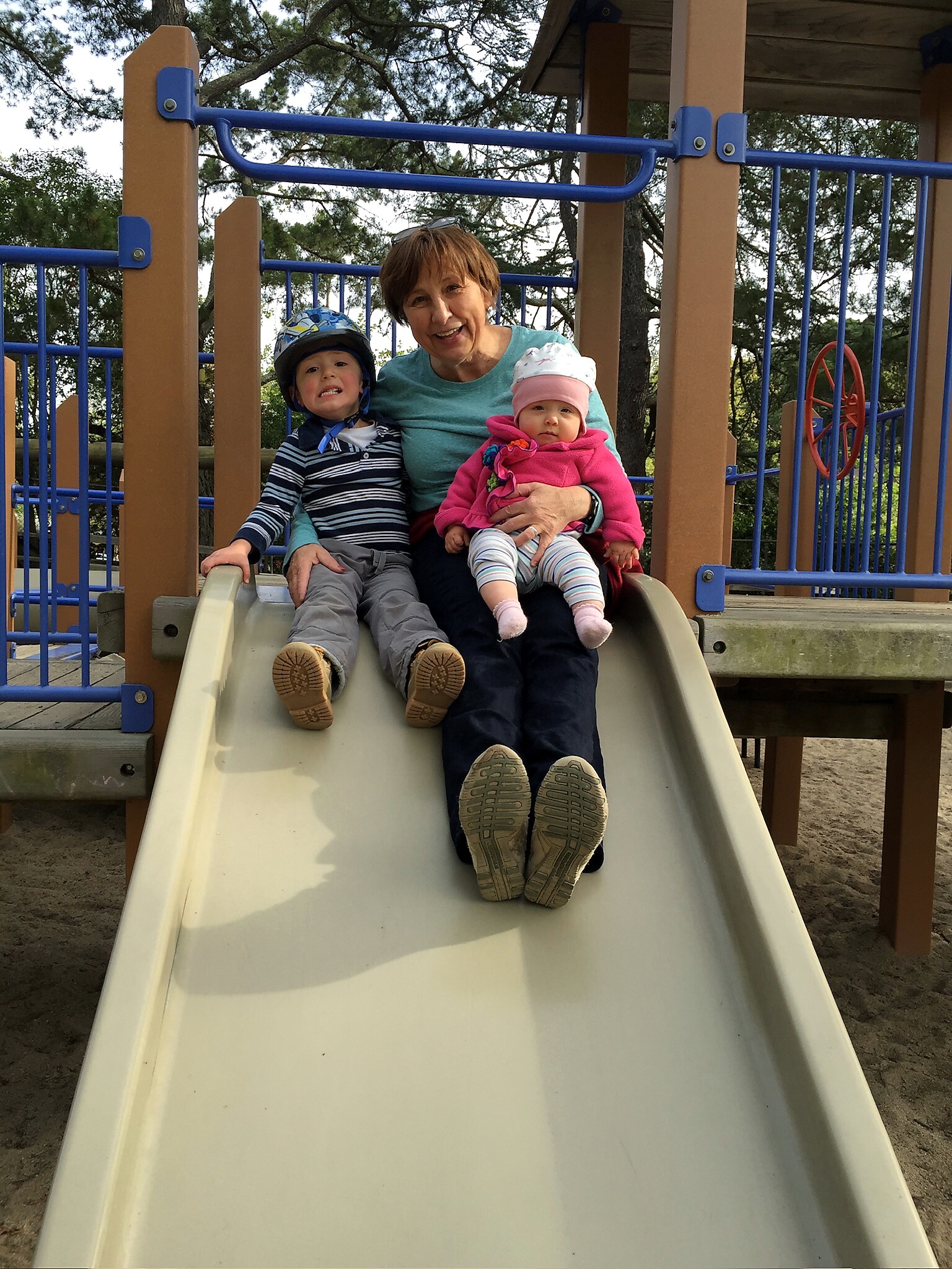 A smiling woman sits on a playground slide with a young boy in a helmet and a baby girl in a pink coat on her lap.