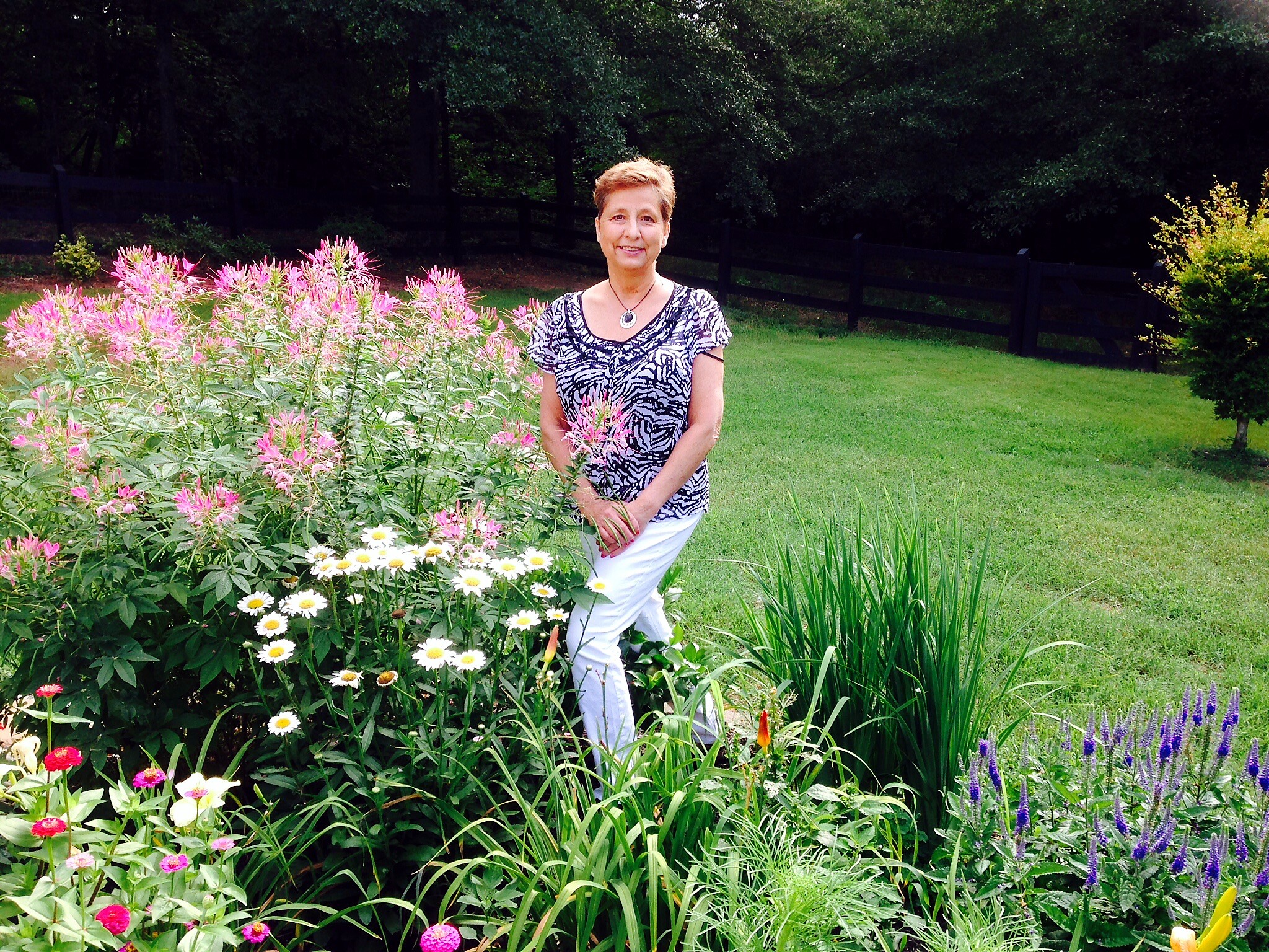 Woman standing in a garden beside pink flowering bushes and white daisies, with a dark fence in the background.