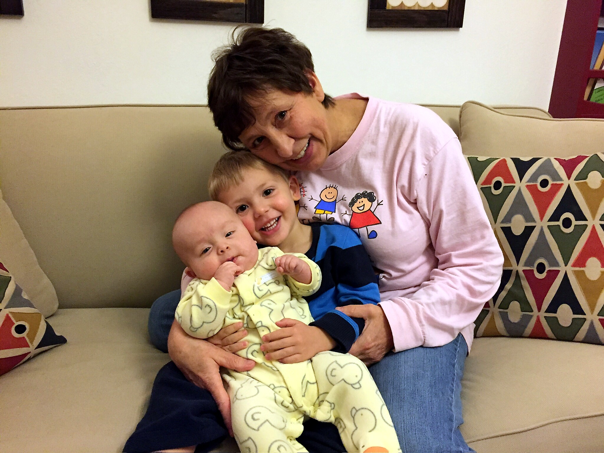 Smiling woman with short dark hair cuddling two babies on a beige couch in a living room.