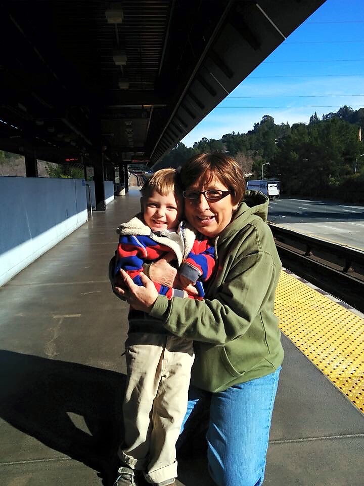 Woman in a green hoodie hugs a smiling boy on an outdoor train platform with yellow tactile paving and tracks nearby.