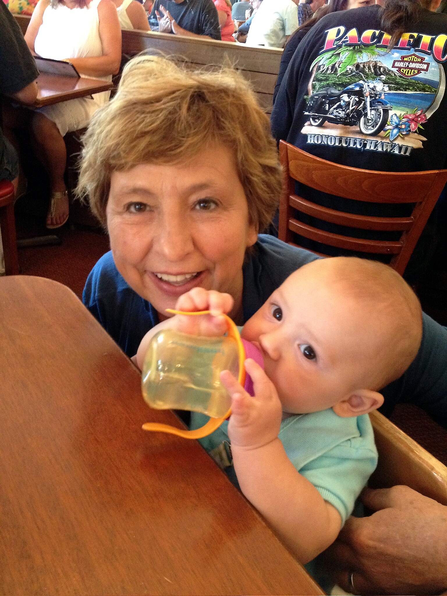 Smiling woman with short blond hair beside a baby drinking from a bottle at a busy restaurant.