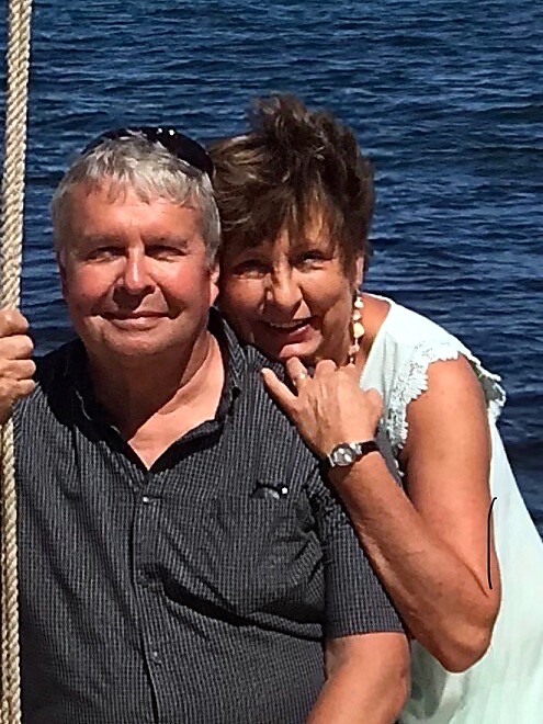 Older couple posing for a photo on a boat by a blue lake, the woman leaning on the man's shoulder with her hand on his chest.