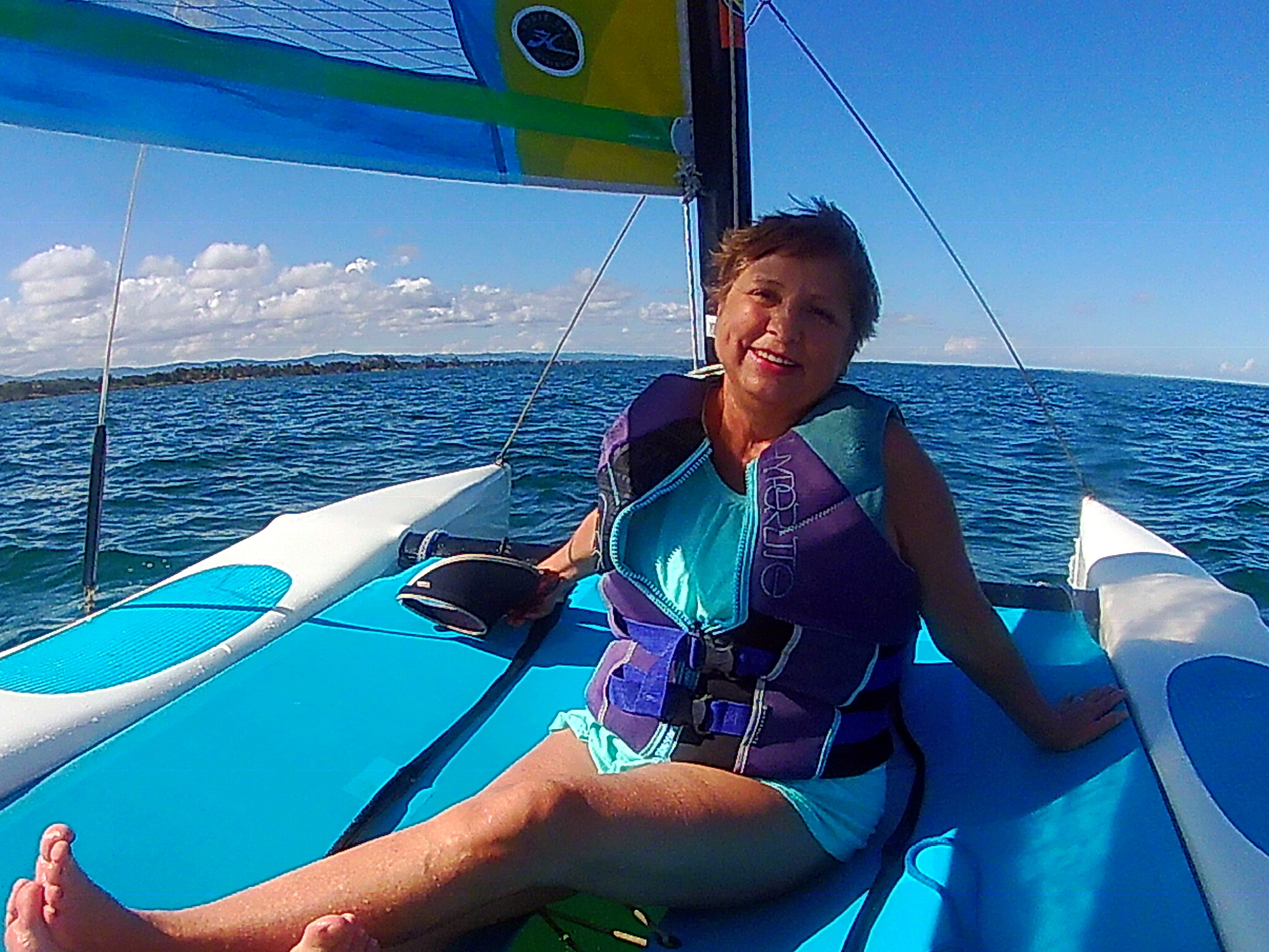 Smiling woman in a purple life jacket seated on a blue sailboat, open water and blue sky behind.