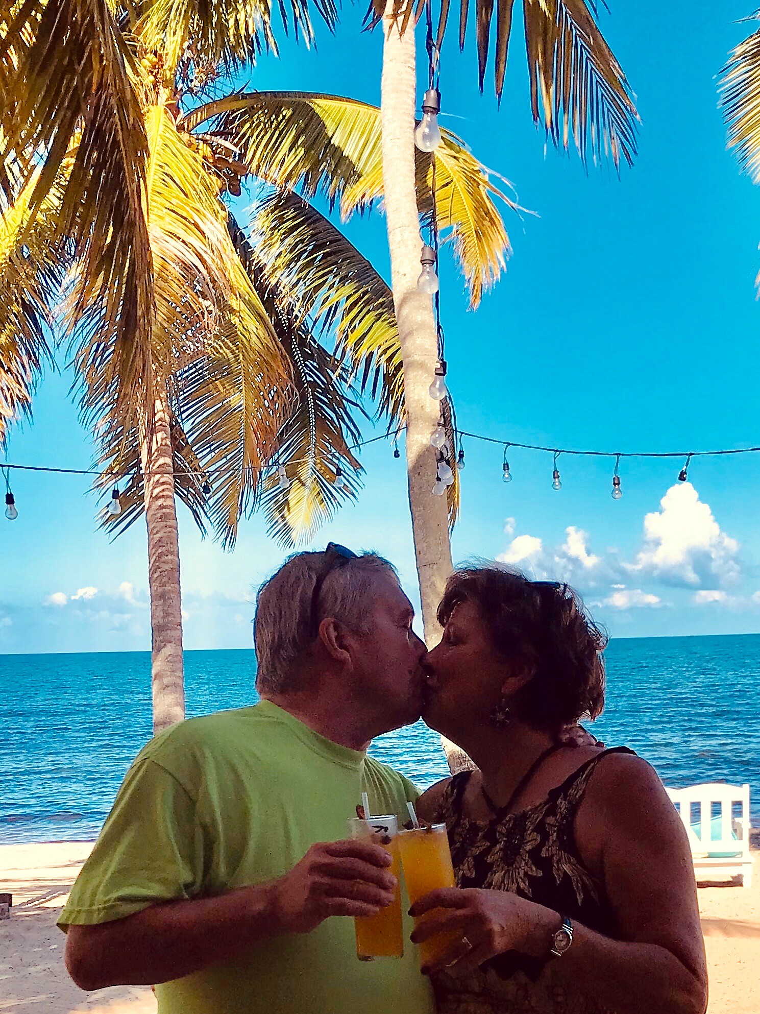 Couple kissing at a beachside bar, holding cocktails with the ocean and palm trees in the background.