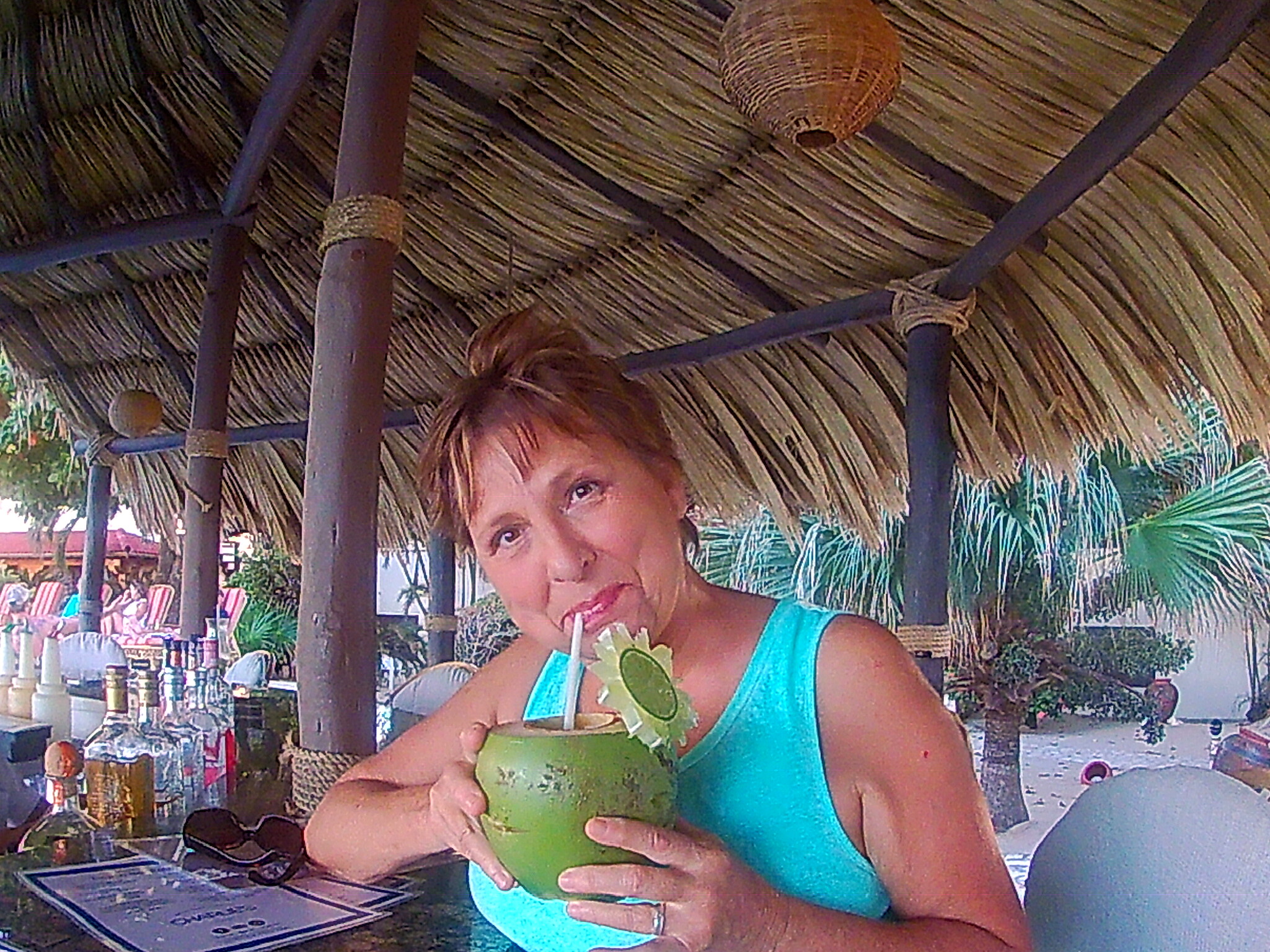 Woman in turquoise top at a beach tiki bar, sipping from a green coconut with a straw.