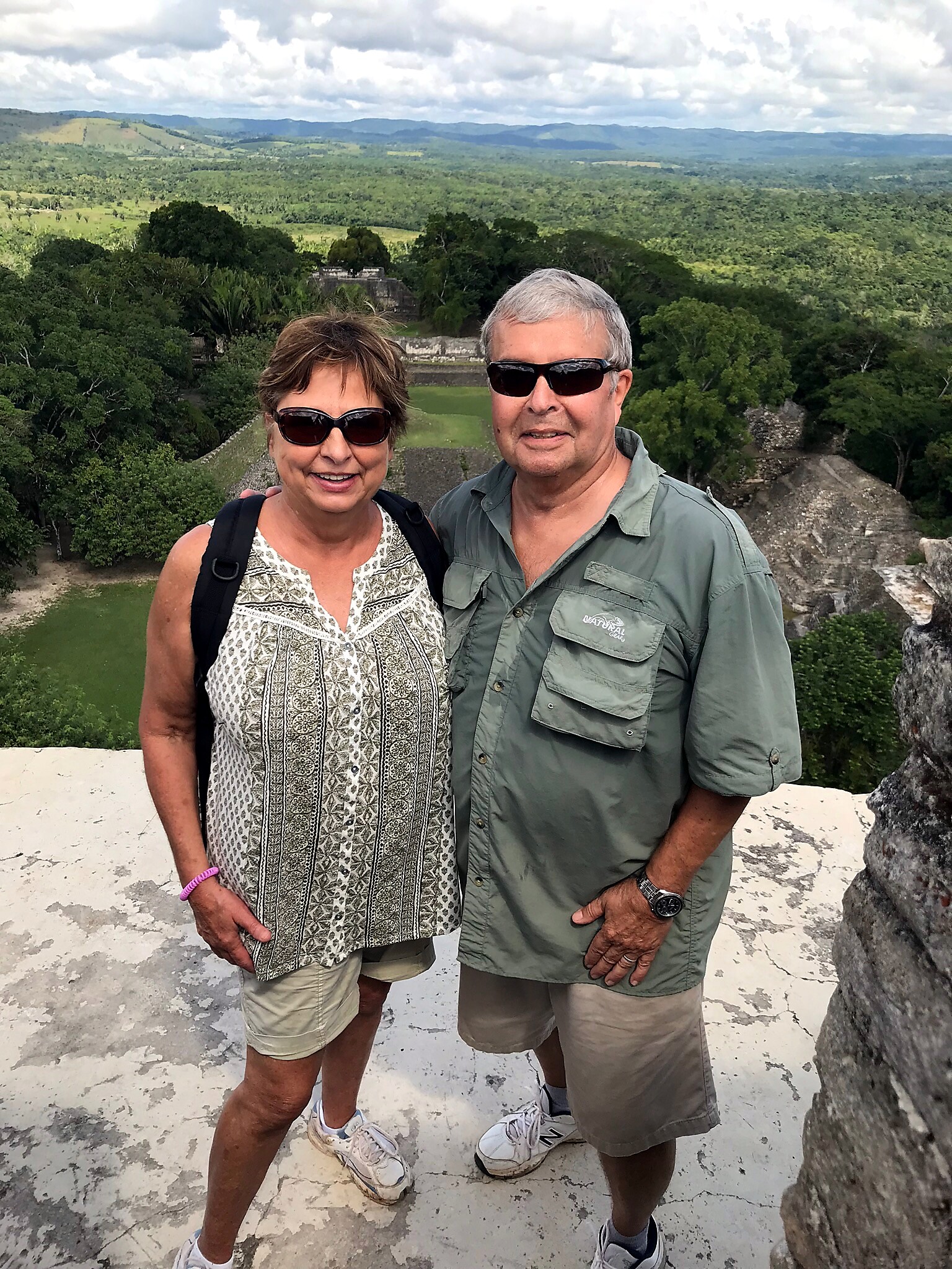 Smiling couple in sunglasses stand together at a stone overlook, with a green valley and distant mountains behind them under a cloudy sky.