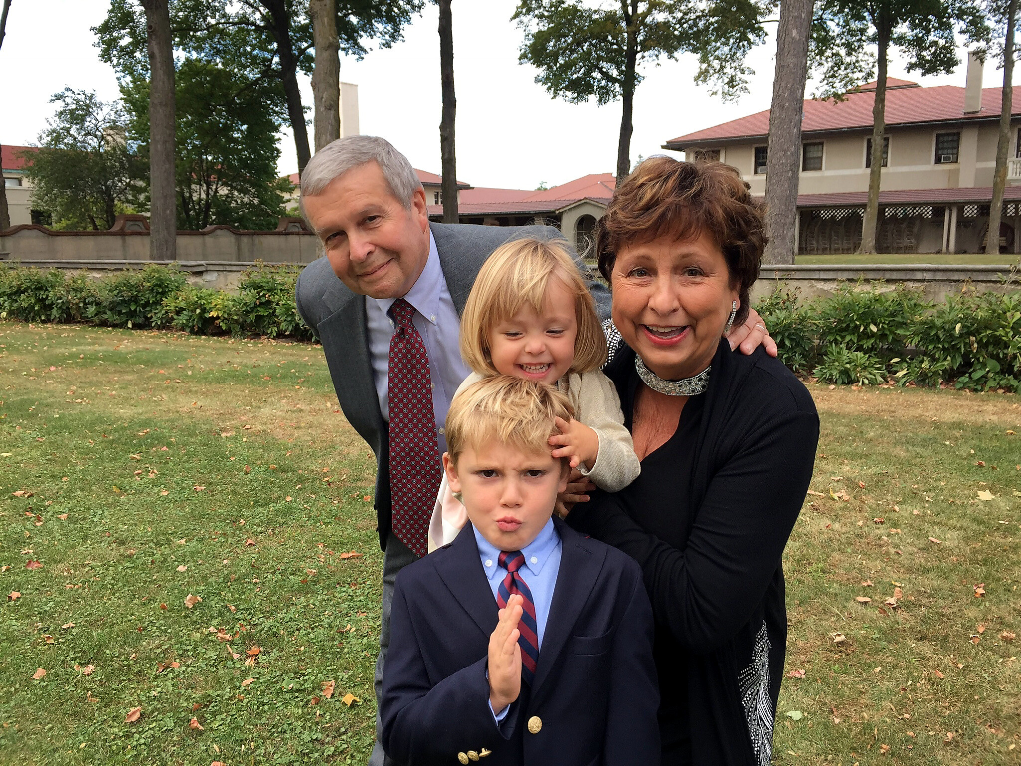 Family of four posing for a photo outdoors in a park, with trees and a building in the background, all smiling.
