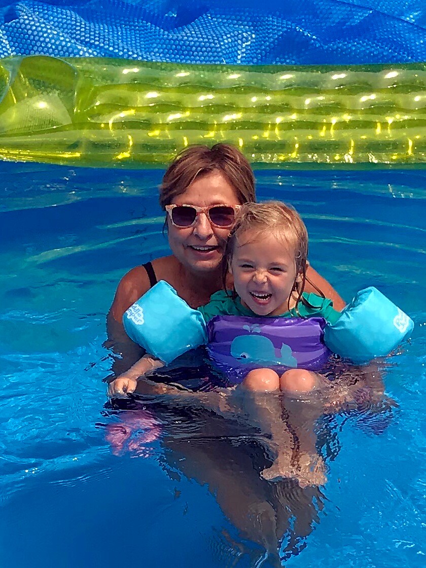 Woman with sunglasses and a child in a purple life jacket and blue arm floats, both smiling in a bright blue pool with a yellow inflatable behind them.