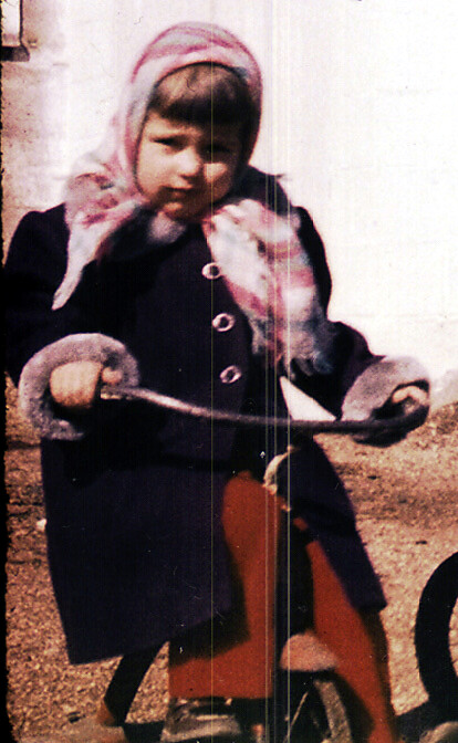 Child in a dark coat and pink scarf sits on a small tricycle outdoors in winter.