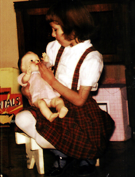 Young girl in a plaid jumper and white blouse sits indoors, holding and tending a pink-dressed doll.