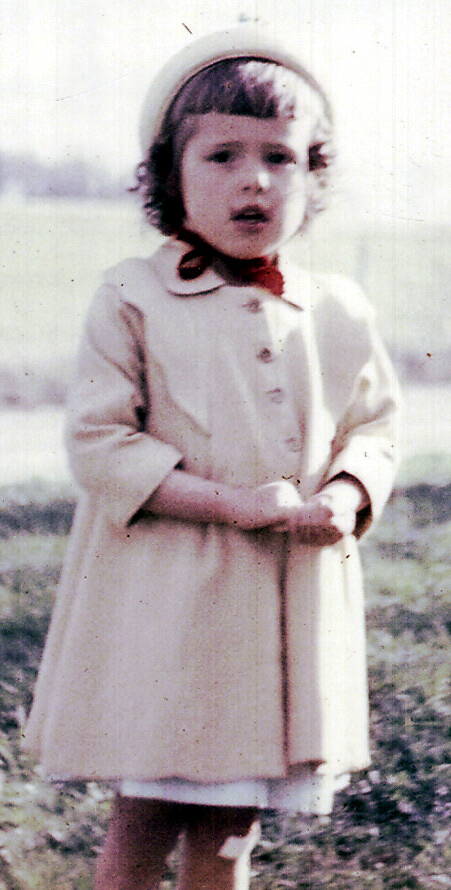 Young girl standing outdoors in a light coat and bonnet, hands clasped, looking at the camera in a grassy setting.
