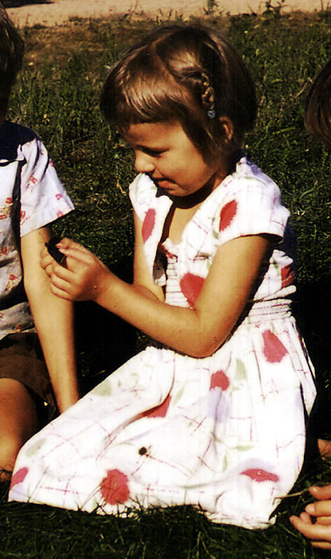 Girl in a white dress with pink patterns sitting on grass outdoors, examining her left arm as a child sits beside her.