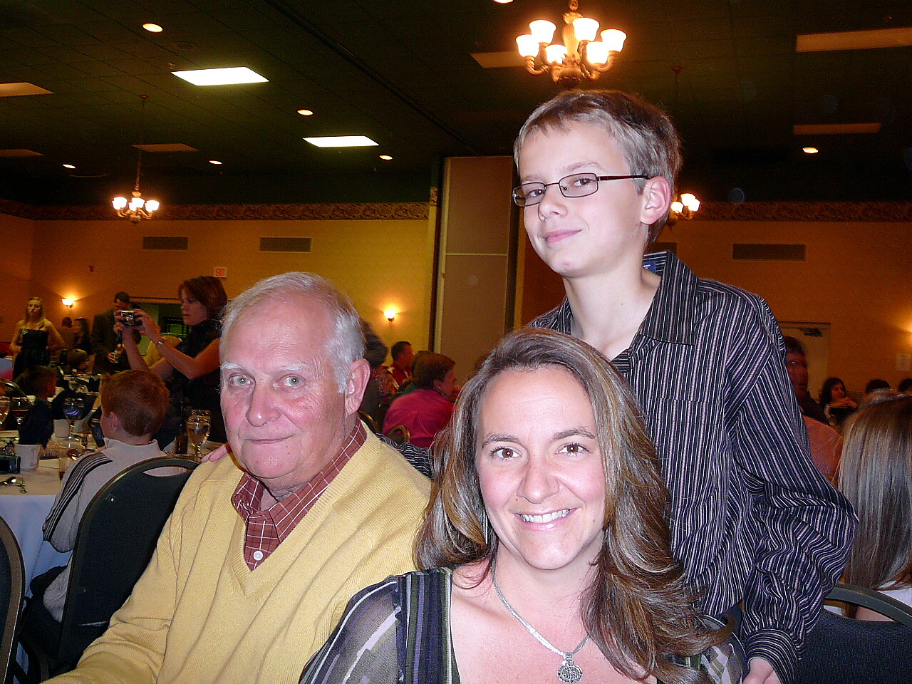 Smiling woman in the foreground with an older man in a yellow sweater and a teenage boy standing behind at a banquet hall dinner table.