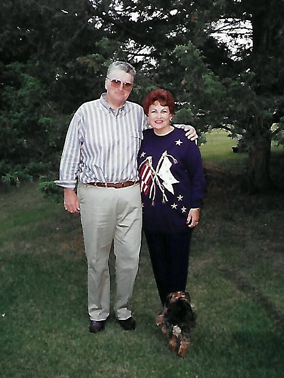 Couple standing on a grassy lawn with trees, man in striped shirt and sunglasses, woman in a dark sweater with star designs, arm around each other; a small dog sits at their feet.
