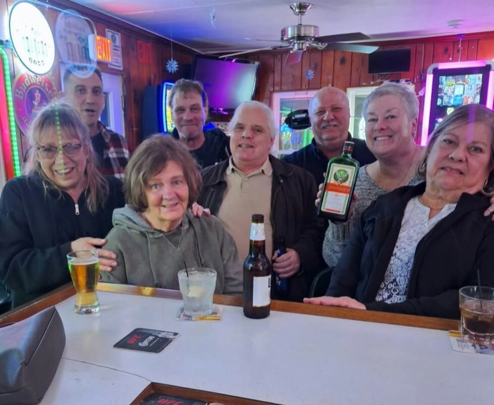 A group of eight adults gathered at a bar, smiling and posing for the photo with drinks on the counter, neon lights in the background.