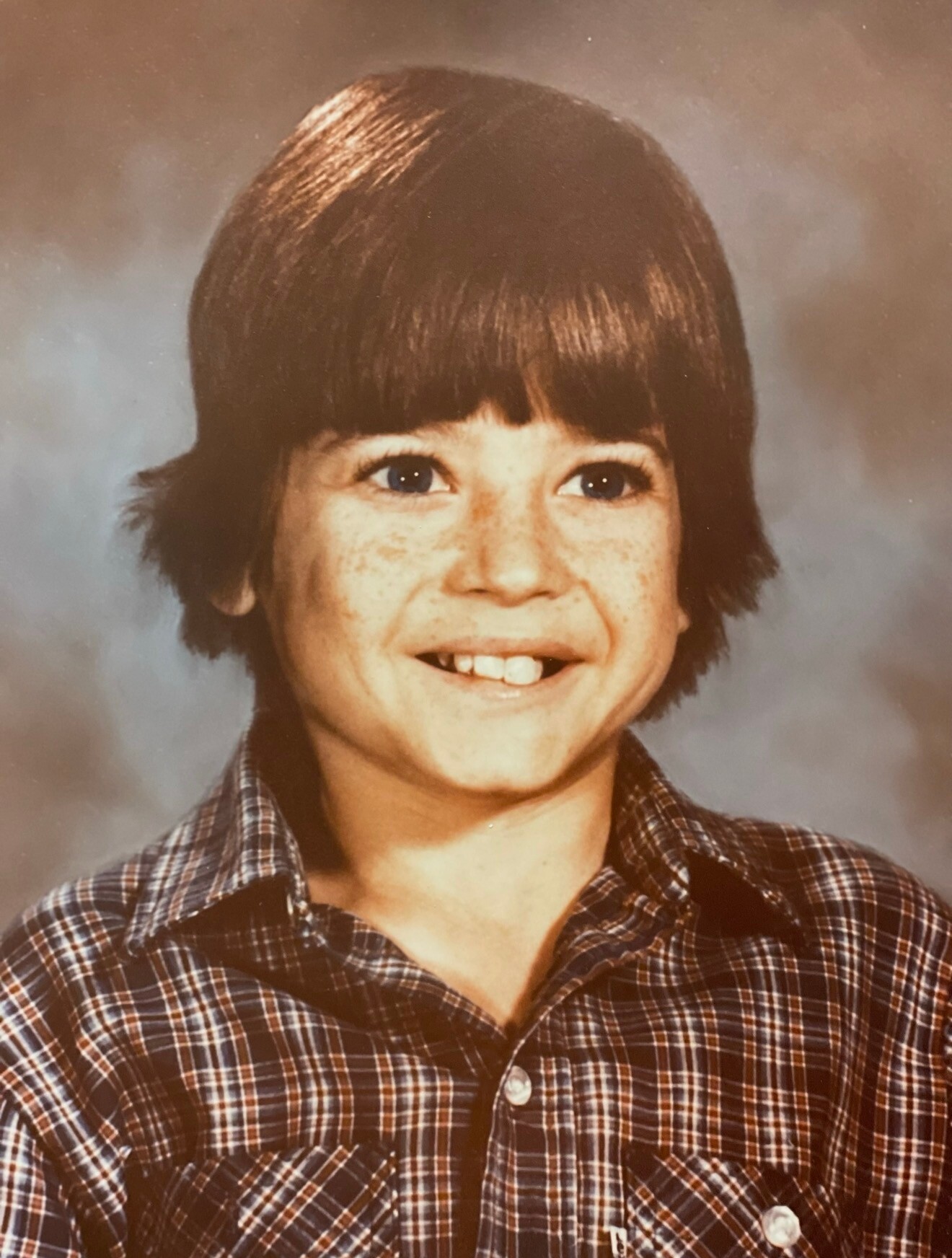 Smiling boy with dark brown hair in a plaid shirt photographed for a school portrait.
