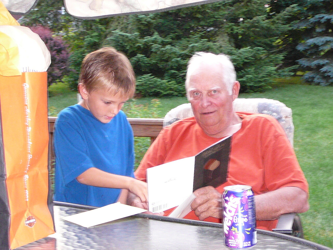 Boy in a blue T-shirt and an older man in an orange shirt sit at a patio table, looking at a card together; a purple can is nearby.