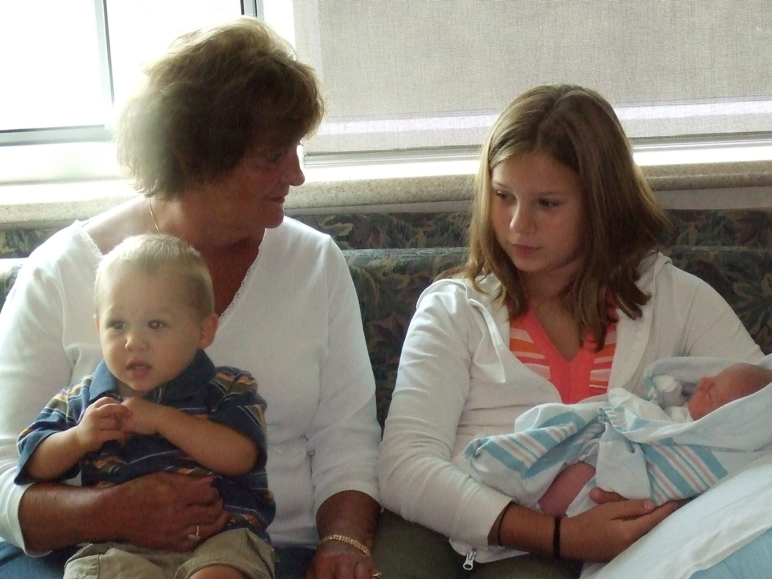 Older woman holding a small baby while a teenage girl sits beside them with a newborn wrapped in a blue striped blanket on her lap and arms nearby.