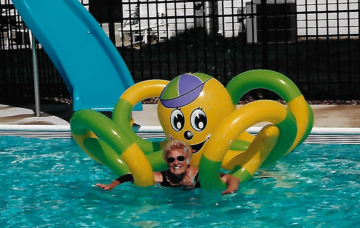 Older woman wearing sunglasses sits in a pool inside a bright yellow-green inflatable octopus with long tentacles, near a blue slide in the background.
