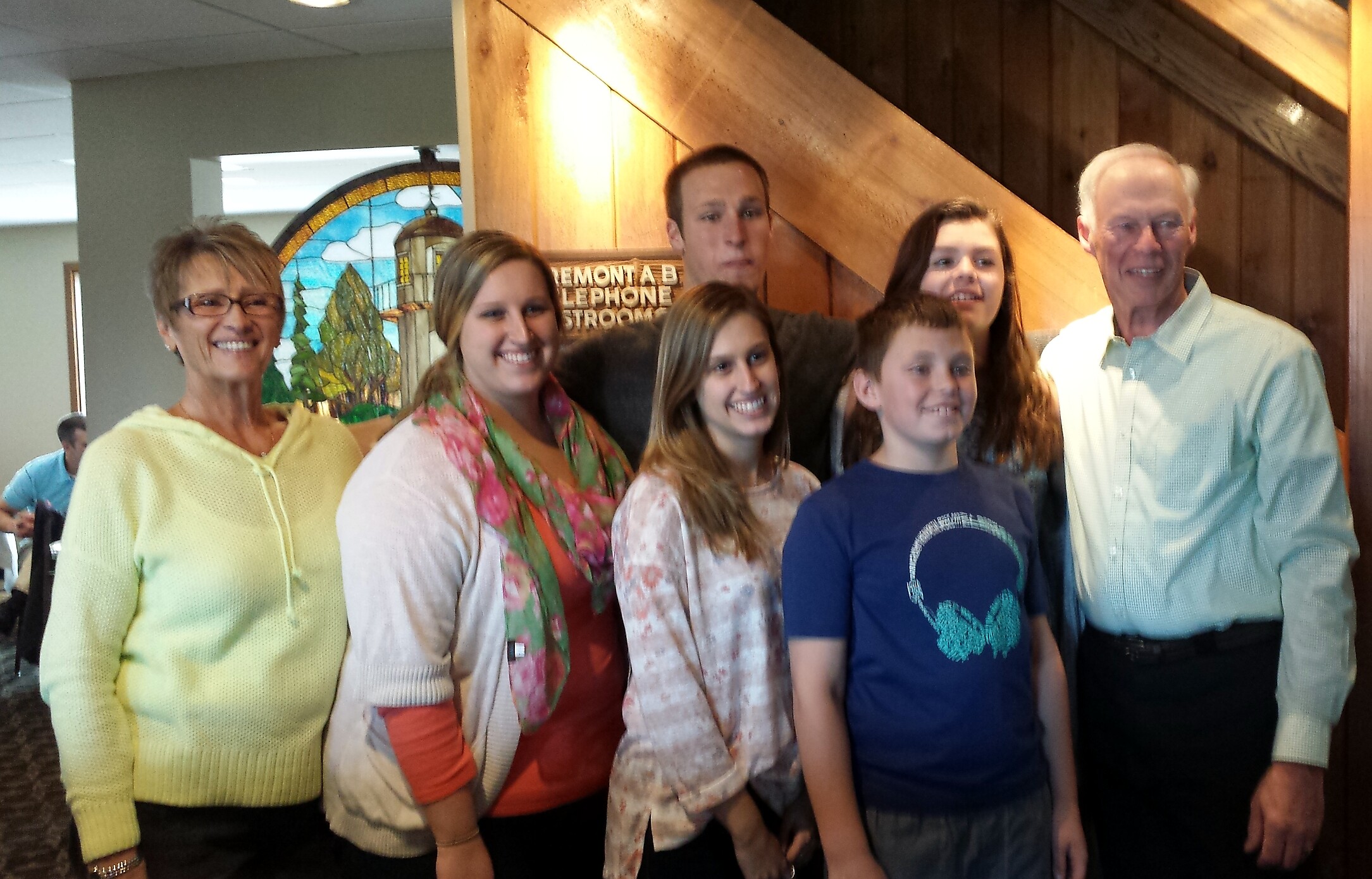 Group of six smiling people posing indoors for a photo, with a stained-glass backdrop and wooden beams behind them.