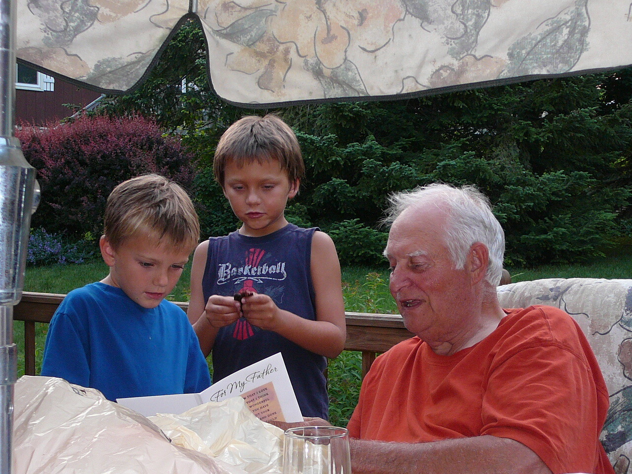 Grandfather in an orange shirt sits on a patio with two young boys, as they look at a card titled 'For My Father' on the table outside under a canopy.