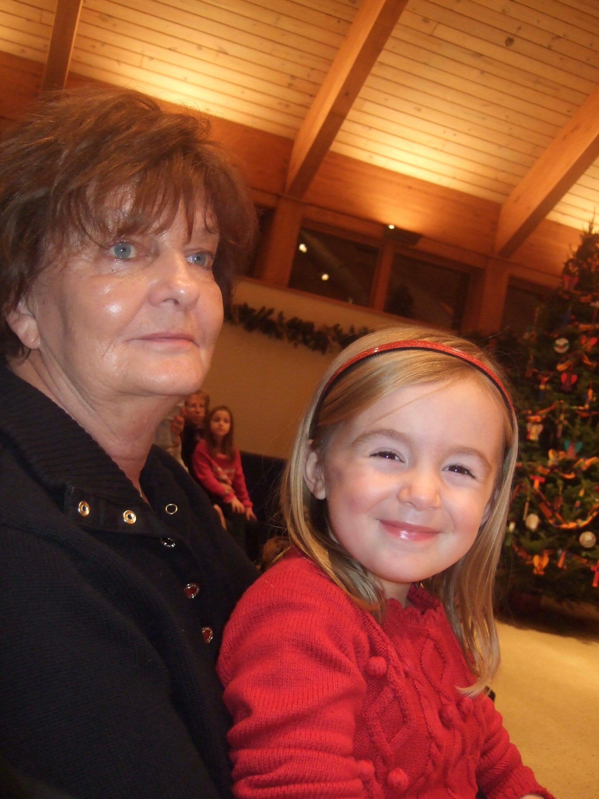 Smiling older woman with short brown hair beside a girl in a red sweater, Christmas tree and warm interior in the background.