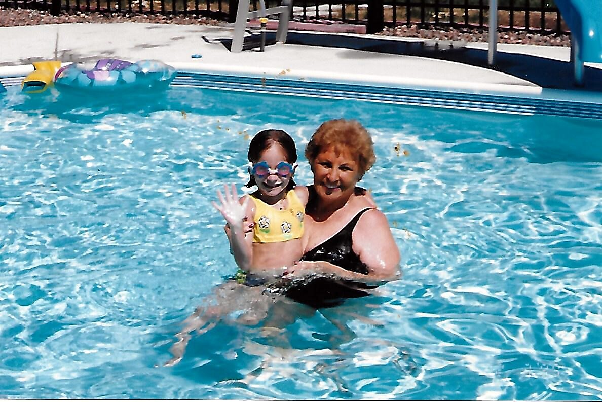 Older woman in a black swimsuit holding a young girl in a yellow swimsuit in a sunny swimming pool, waving at the camera.
