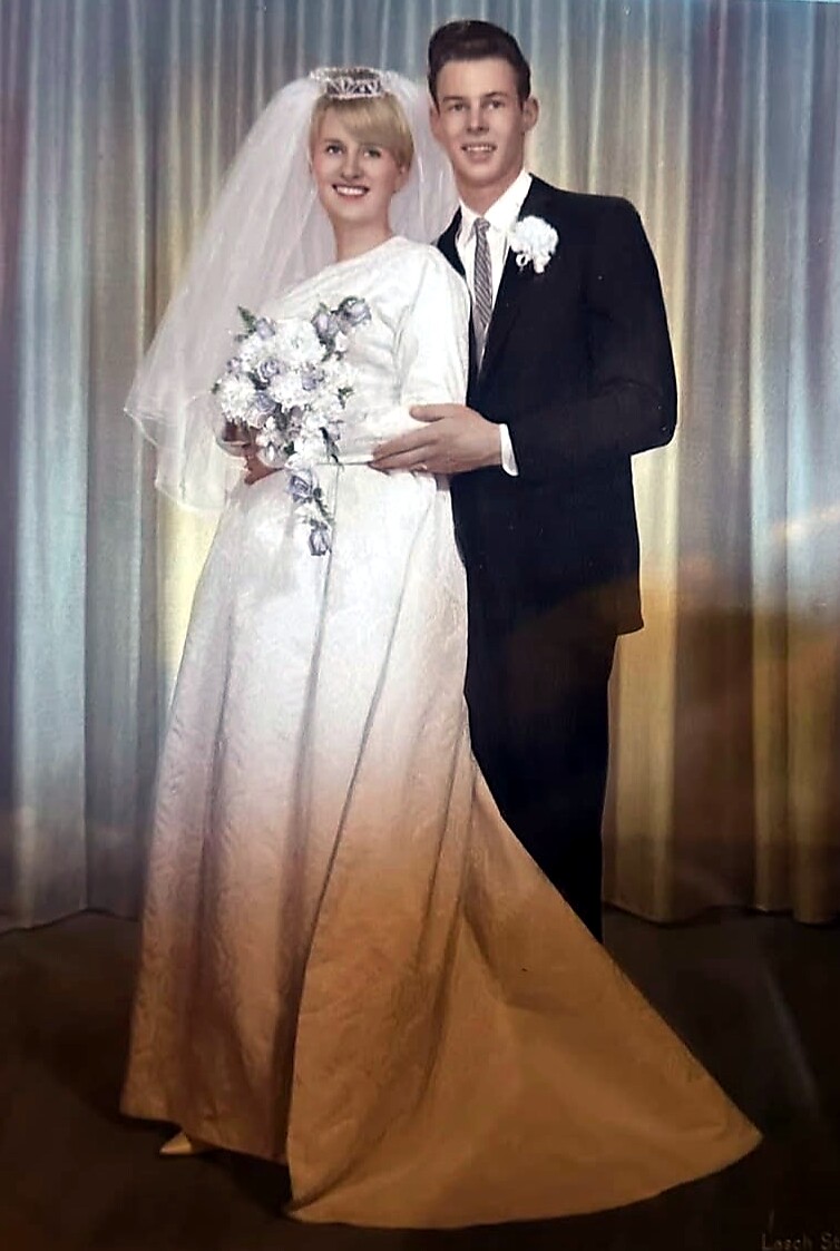 Bride and groom posing together on their wedding day; bride in white gown with veil and bouquet, groom in dark suit with boutonniere, standing in front of a curtain backdrop.