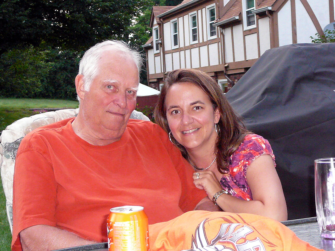 A smiling older man in an orange shirt sits beside a woman in a colorful top outdoors, with a grill cover and a house in the background.