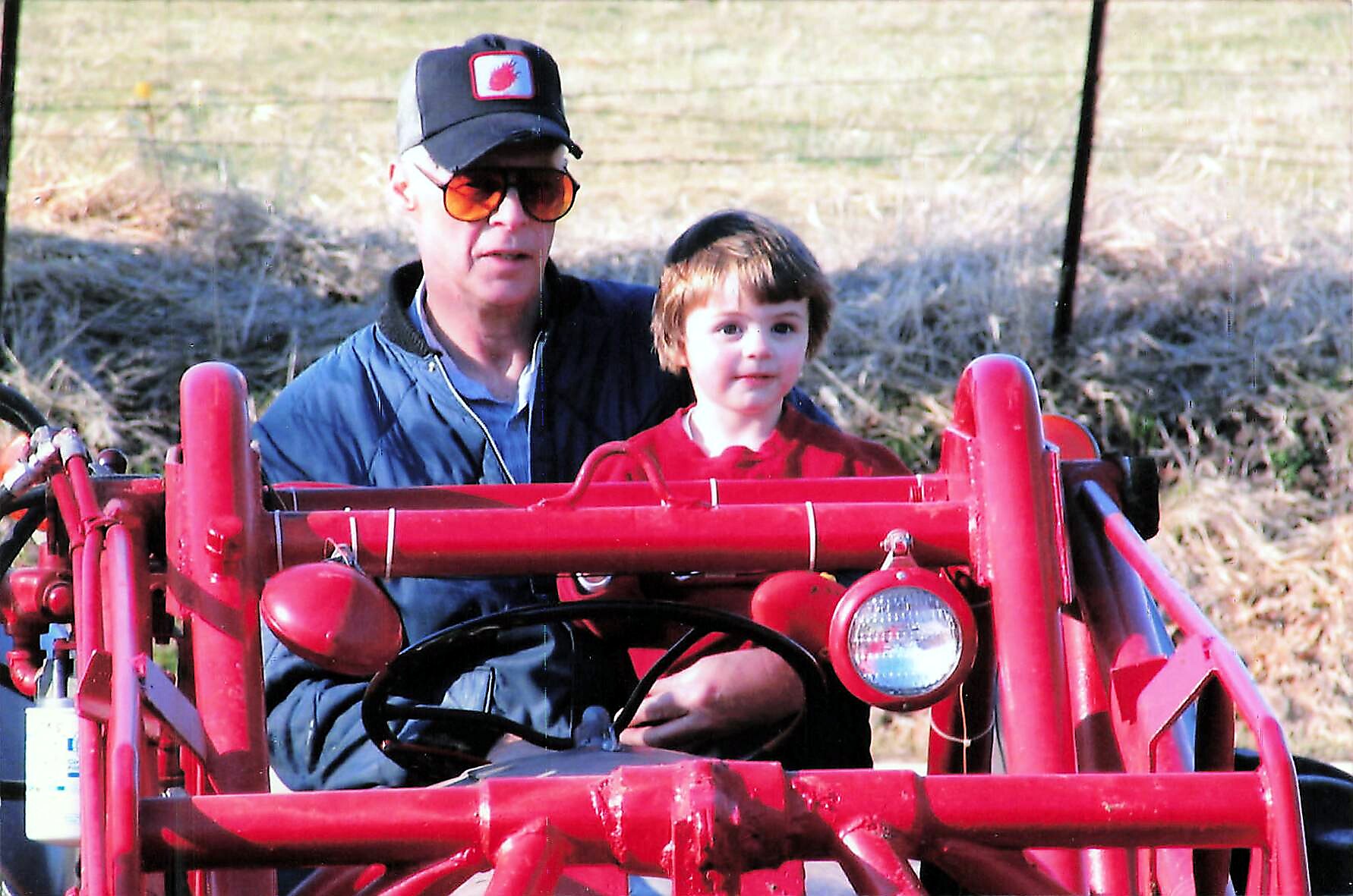 Man in a cap and orange sunglasses sits with a young child on a bright red tractor outdoors in a field.