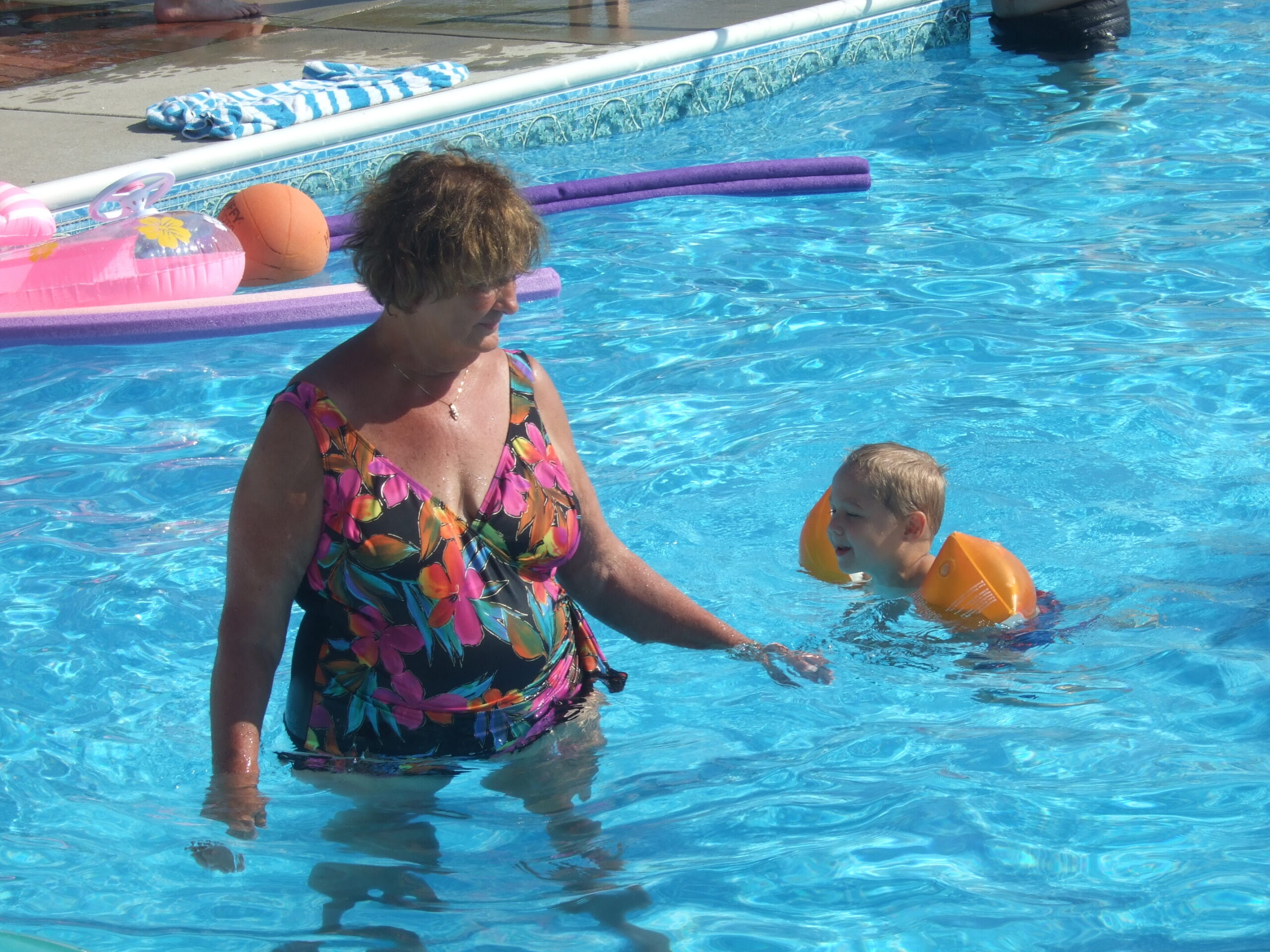 Woman in a colorful swimsuit standing in a sunny blue pool near inflatable toys and a child with orange arm floaties nearby.