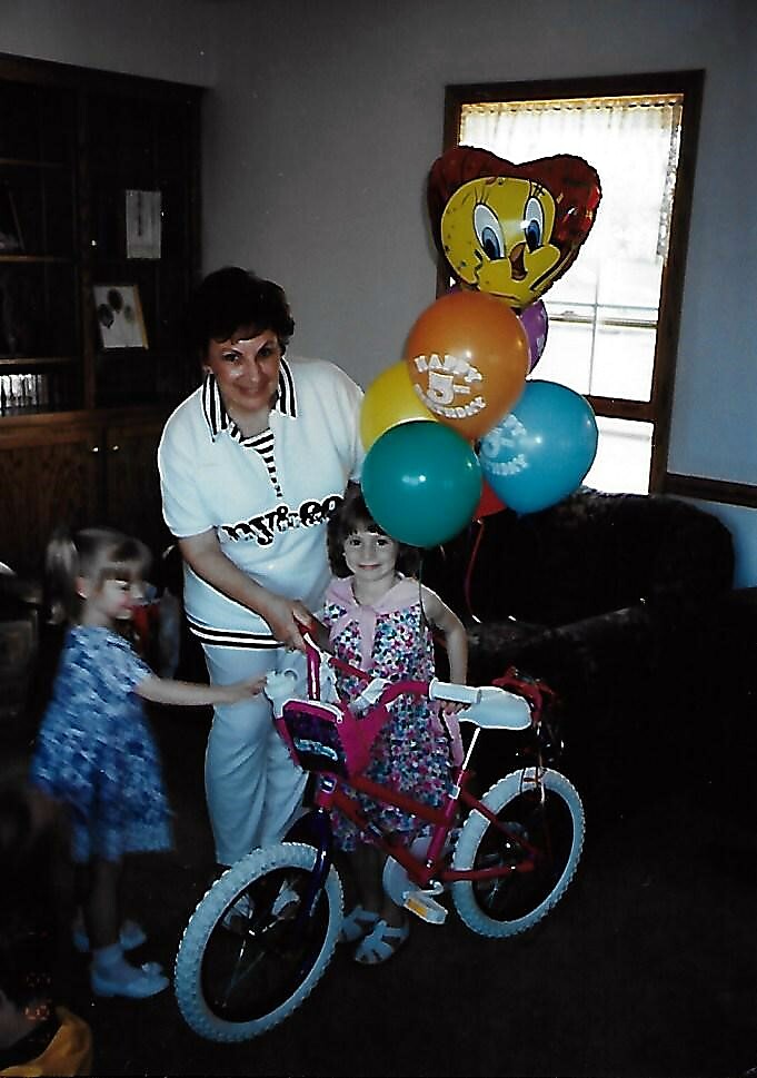 Birthday party scene: a woman helps a young girl with a pink bicycle, balloons tied above in a home setting.