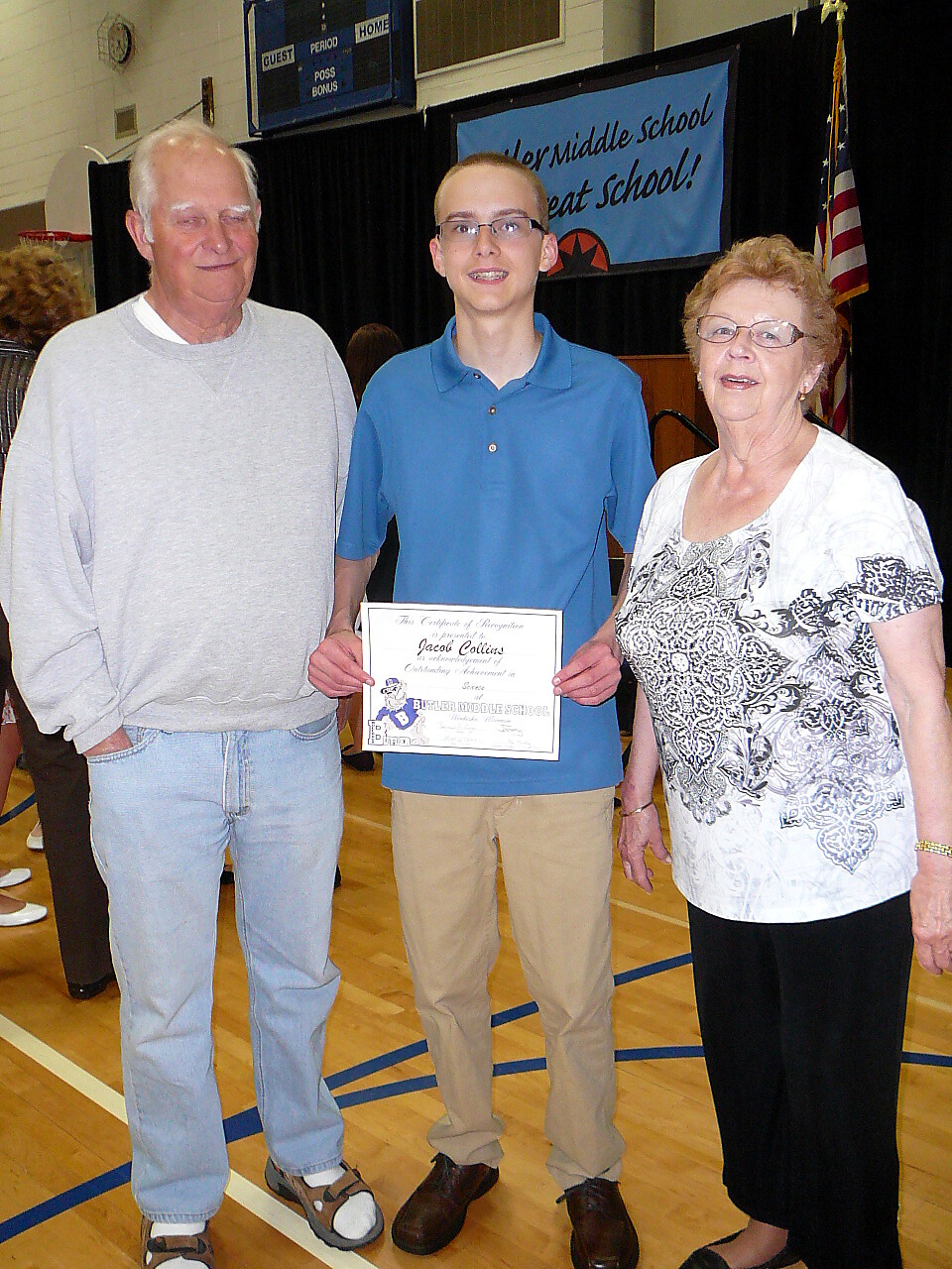 Three people stand in a gym during an award ceremony: an older man in a gray sweatshirt, a smiling student in a blue polo holding a certificate, and an older woman in a white patterned blouse.