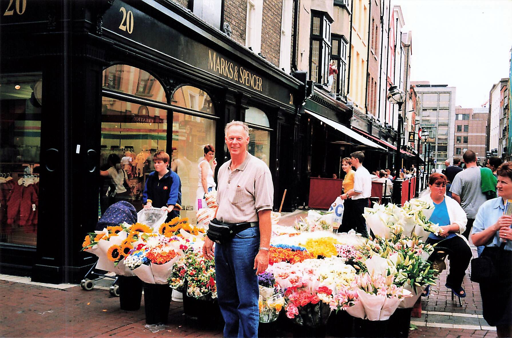 Flower stall along a shopfront street, colorful bouquets arranged outside a Marks & Spencer store, busy with pedestrians nearby.