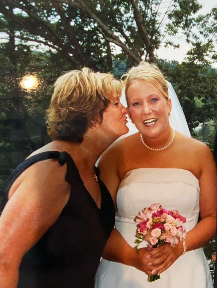 Bride in a strapless white wedding dress holding a pink bouquet, smiling while a guest in a black dress kisses her cheek outdoors.